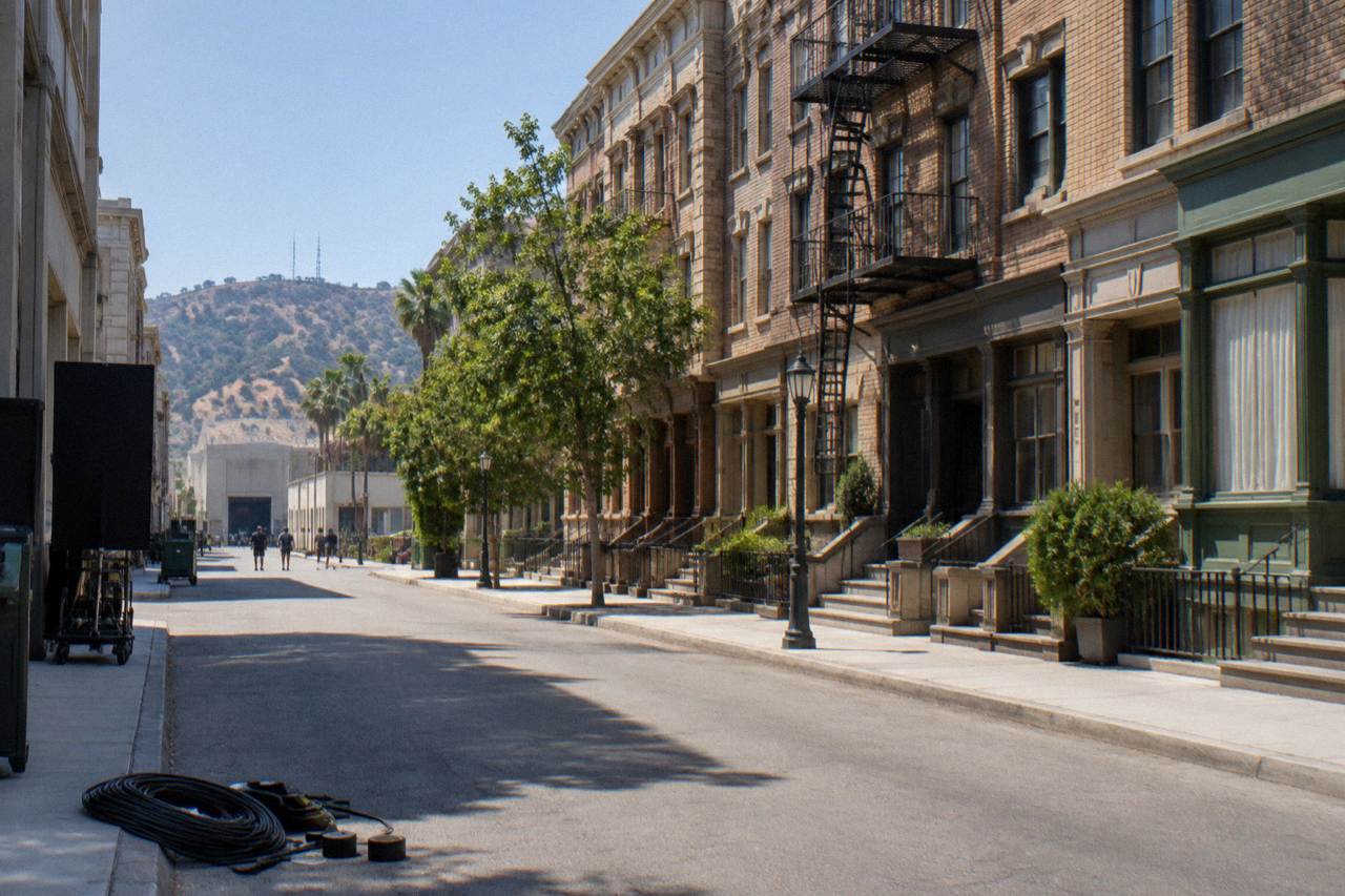 Sunlit Warner Bros. backlot street facade with brownstone storefronts and a quiet production lot road