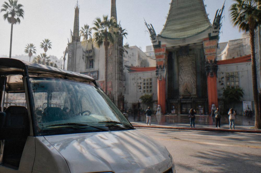 A guided tour van pulled over at a curb while passengers photograph a historic theater facade
