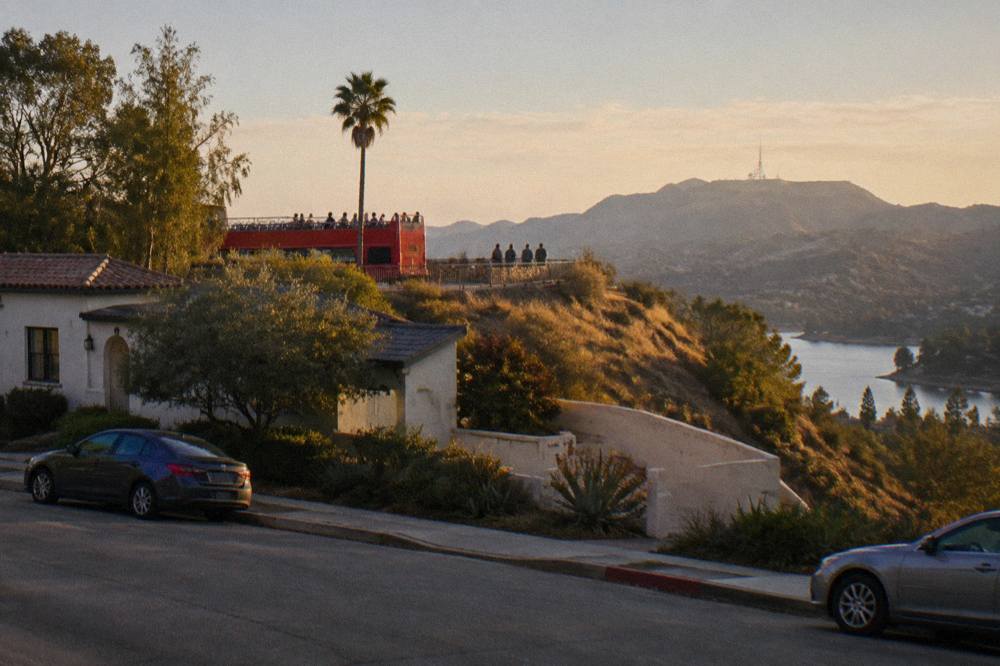 An open-top tour bus on a Hollywood Hills street with the sign visible on the ridge beyond