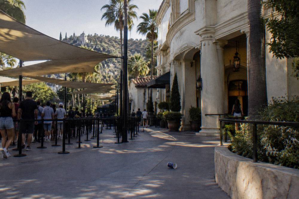 Theme park midway with a show palace facade and queues winding beneath shade structures