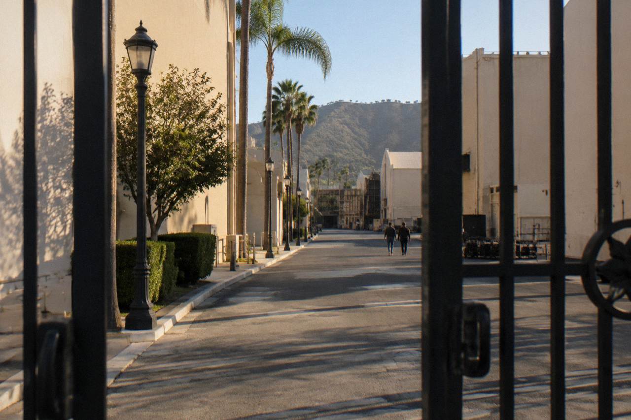A gated studio backlot street with soundstage walls and lampposts under a clear California sky